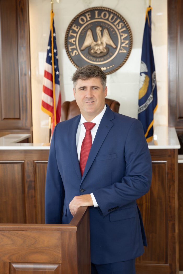 An injury lawyer in a navy suit and red tie stands in front of the Louisiana state seal and flags.