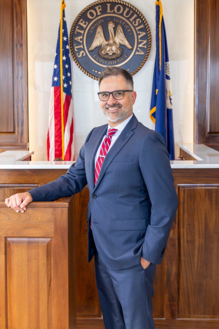 An injury lawyer in a suit and glasses stands before the Louisiana state seal and flags.