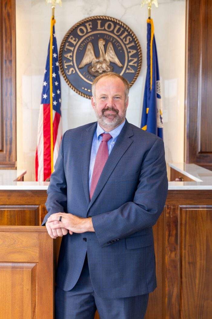 An injury lawyer in a suit and tie stands in front of the Seal of the State of Louisiana and two flags.