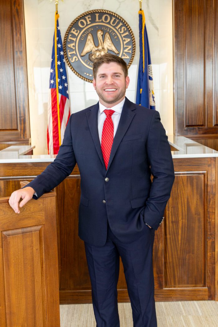 An injury lawyer in a suit and red tie stands in a courtroom in front of the Louisiana state seal.