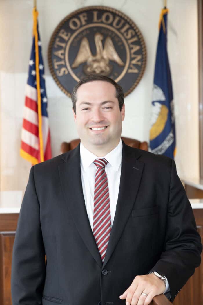 An injury lawyer in a suit and striped tie standing in front of the Louisiana state seal and two flags.