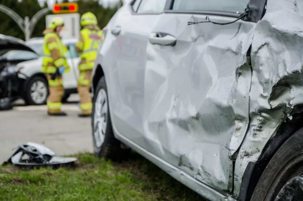 A silver car with significant side-panel damage at an accident scene where an injury lawyer might be needed, with two firefighters in the background.