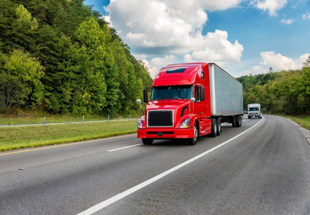 A red semi-truck driving on a highway with a white trailer, followed by a smaller truck in the distance.